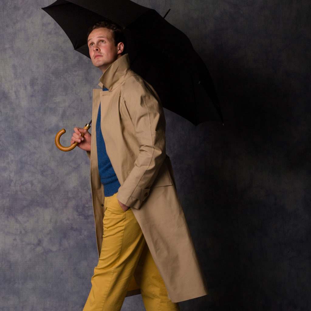 A man walking along in a tan Cordings Duncan Mackintosh holding a black umbrella
