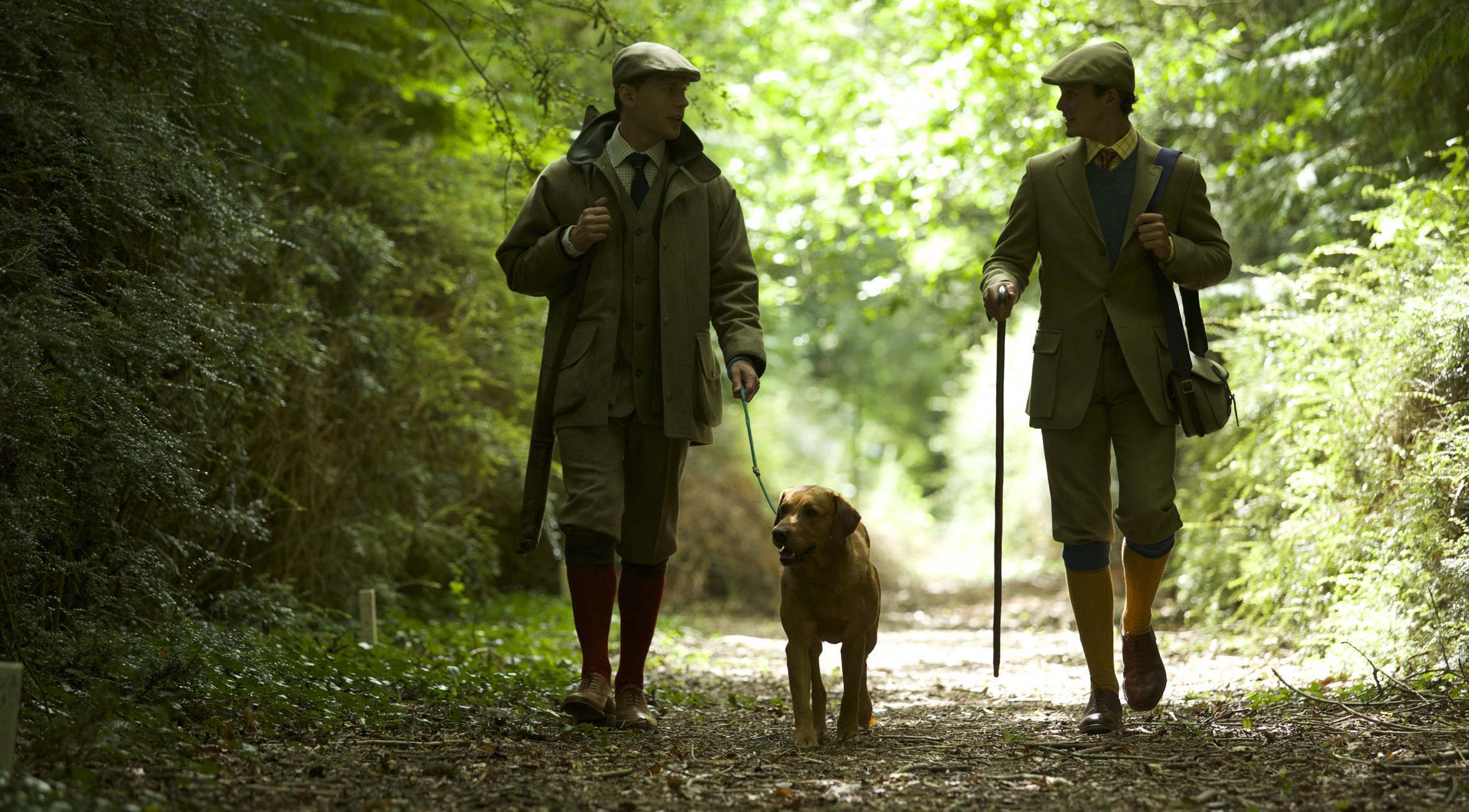 Two men dressed in green tweed shooting clothing walking along a forest pathway with a golden Labrador