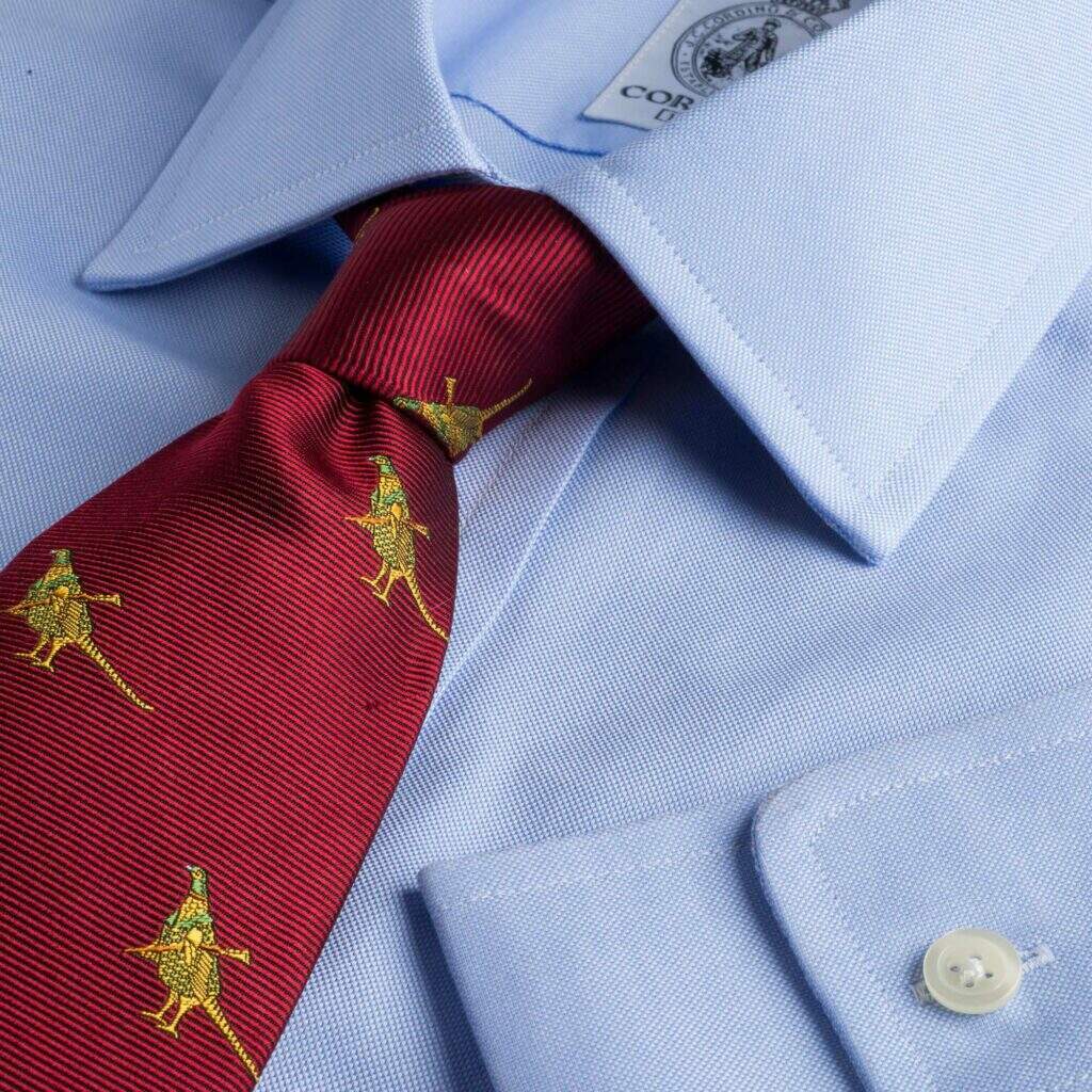 Image of a classic blue oxford shirt, paired with a rich maroon tie that is decorated with pheasant detailing