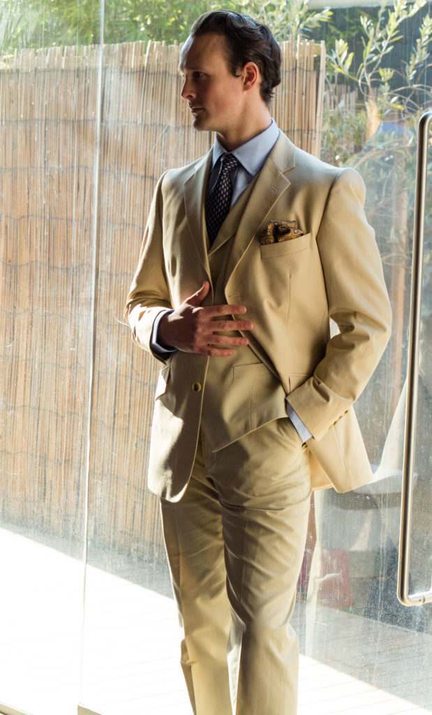 Man dressed in Great Gatsby style stood next to a window, wearing a lightly coloured three piece suit, with a pale blue shirt, a dark blue tie, and a gold pocket square