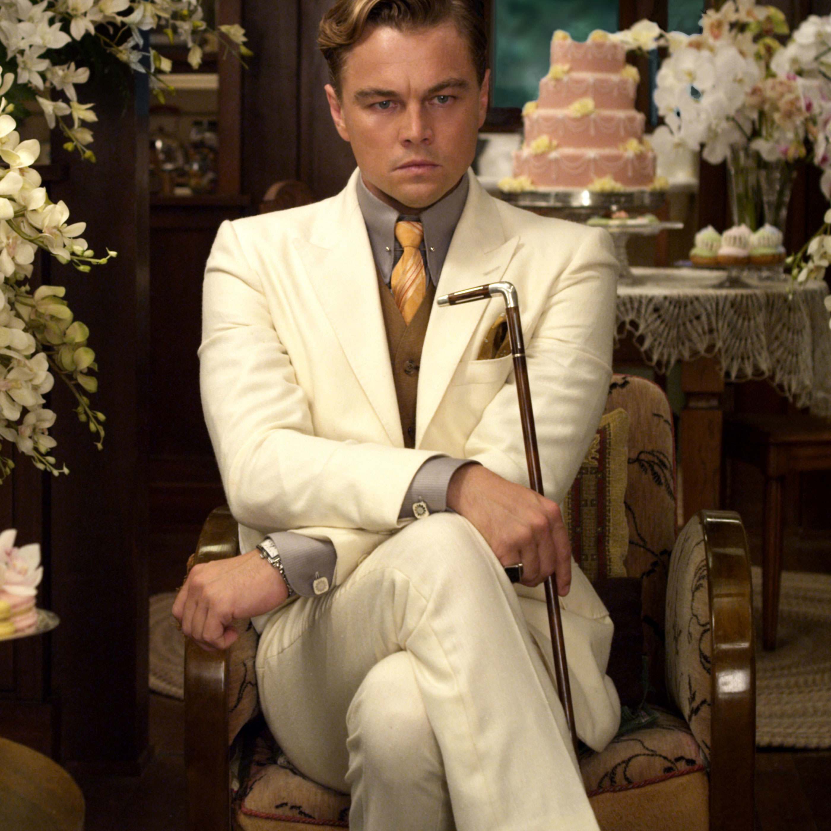 An image of Great Gatsby, played by Leonardo Di Caprio, from the 2013 film adaptation, sat in a white suit with a grey shirt and yellow tie, holding a cane