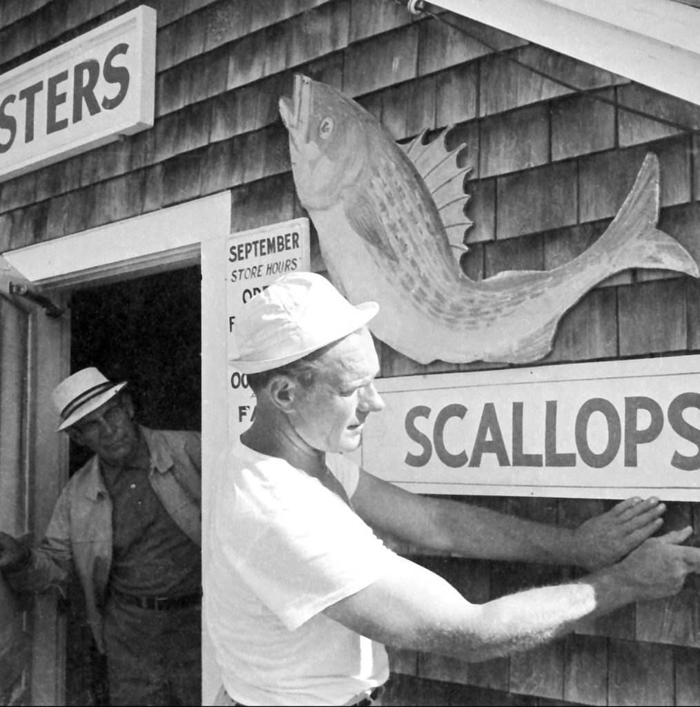 Two men in white clothing pictured working outside a seafood shack selling scallops.