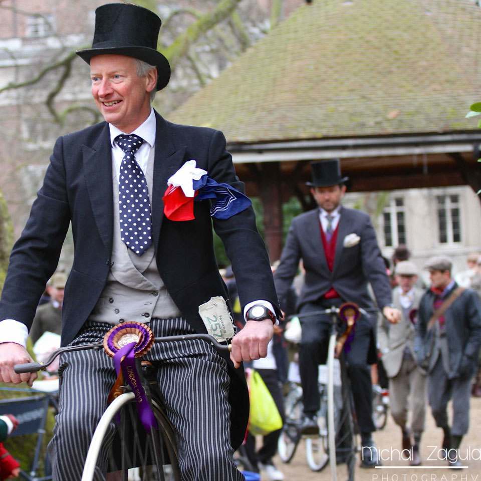 A gentleman in a navy blue tweed suit and top hat riding a bicycle at the Tweed Run in London
