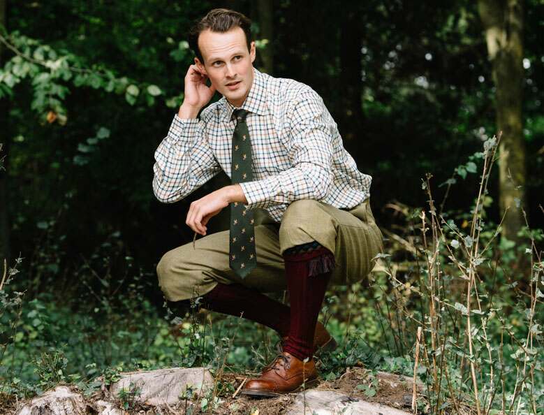 Man crouched on a tree stump wearing brown leather shoes, dark red shooting stockings, tweed breeks and a checked shirt with green tie.