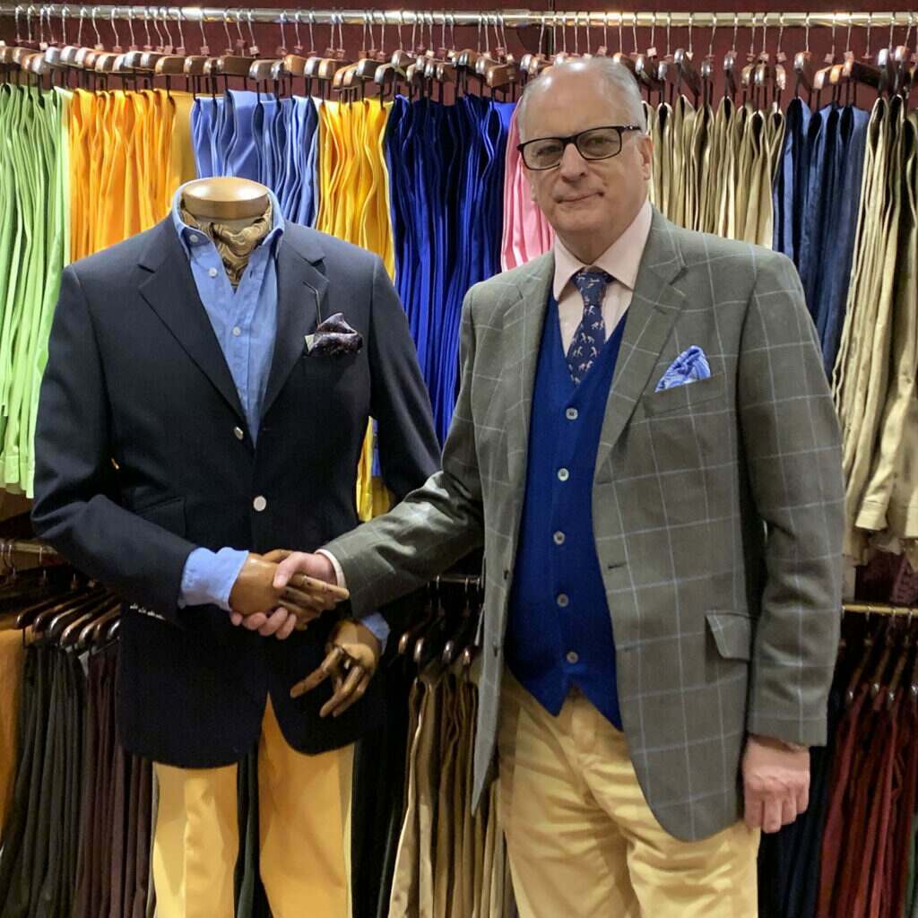 A cordings shop assistant dressed in a dark green jacket, royal blue waistcoat, pink shirt and yellow trousers shaking the wooden hand of a mannequin