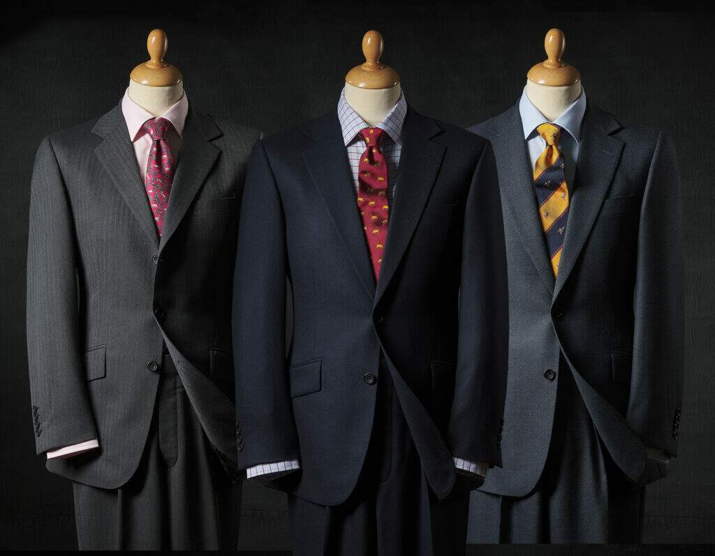 Three mannequins in suits, one in a pinstripe charcoal suit with pink shirt and cherry tie, one in a dark navy suit with a blue and red tattersall shirt and a red tie, and the final mannequin wearing a dark grey suit with a pale blue shirt and navy and yellow tie